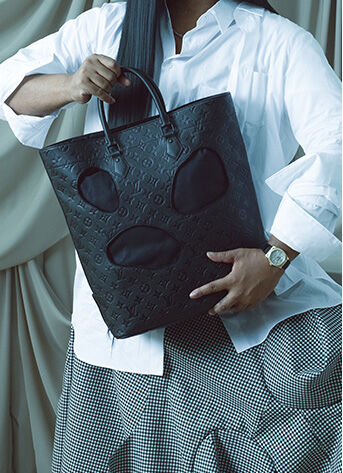Collector's Circle: Law Roach holding a Louis Vuitton Comme Des GAR&Ccedil;ONS x Louis Vuitton Black Monogram Empreinte Bag with Holes in front of beige curtains 