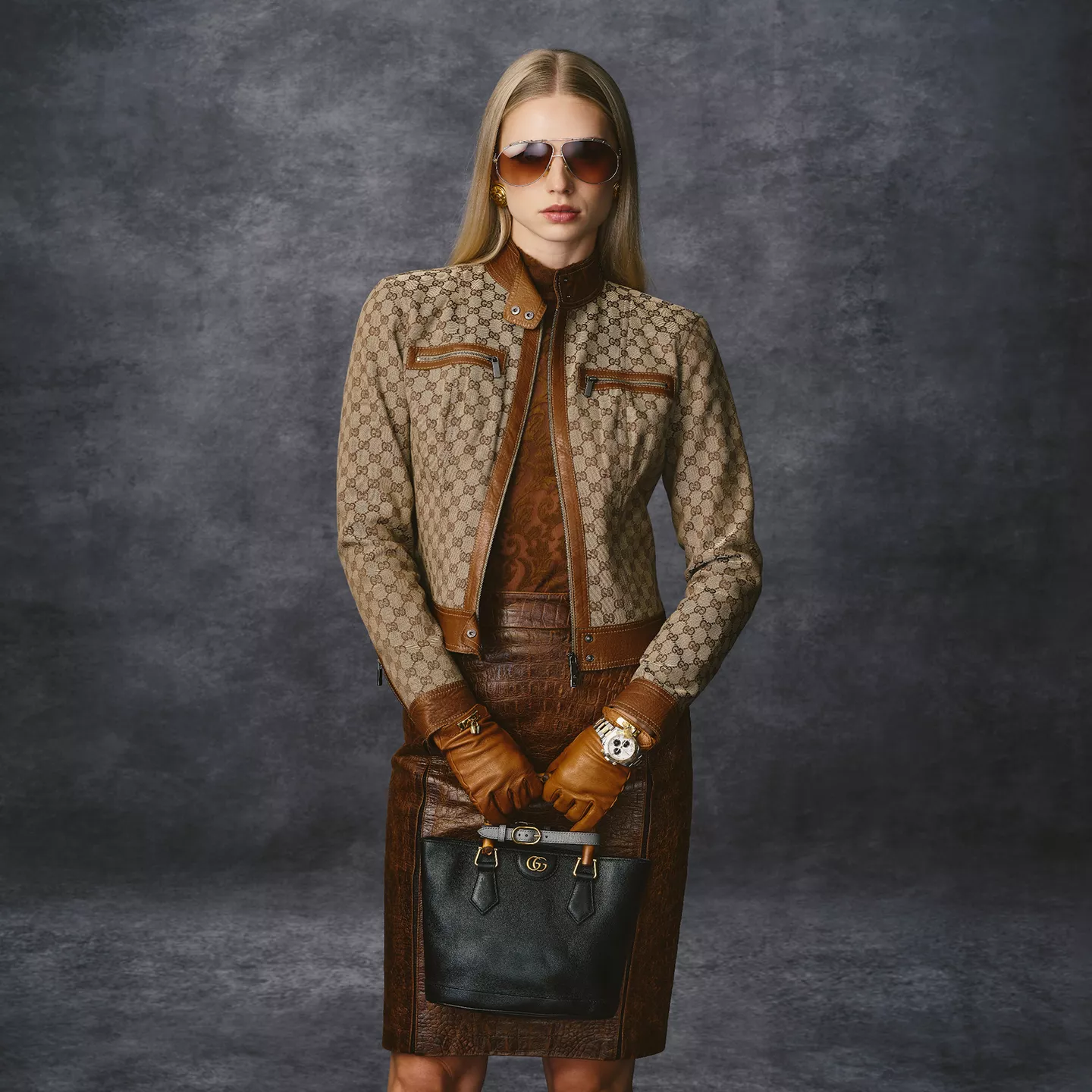 202609_Gucci Feature: A model wearing a monogram Gucci canvas moto jacket, a brown shirt and skirt, Hermes brown leather gloves, and a Gucci black bamboo top handle bag.