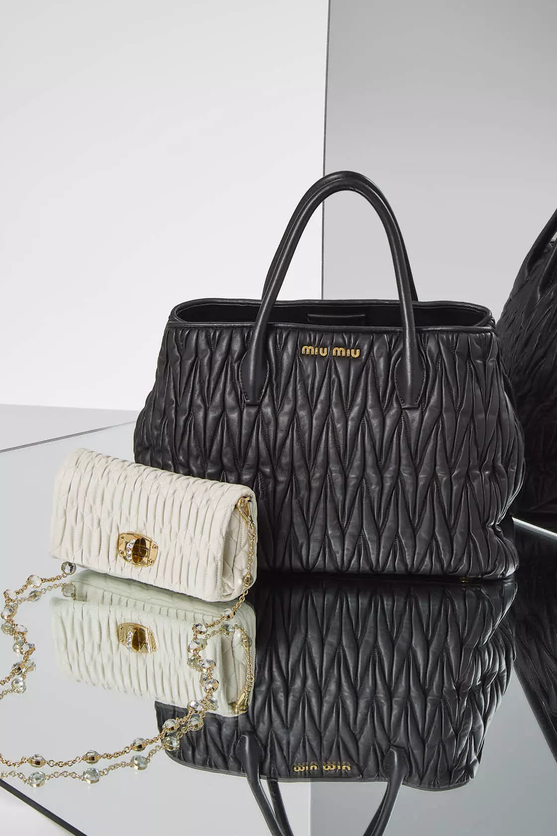 Shop by Brand: Miu Miu White leather nappa WOC in front of a Miu Miu black leather top handle tote bag on a mirrored surface.
