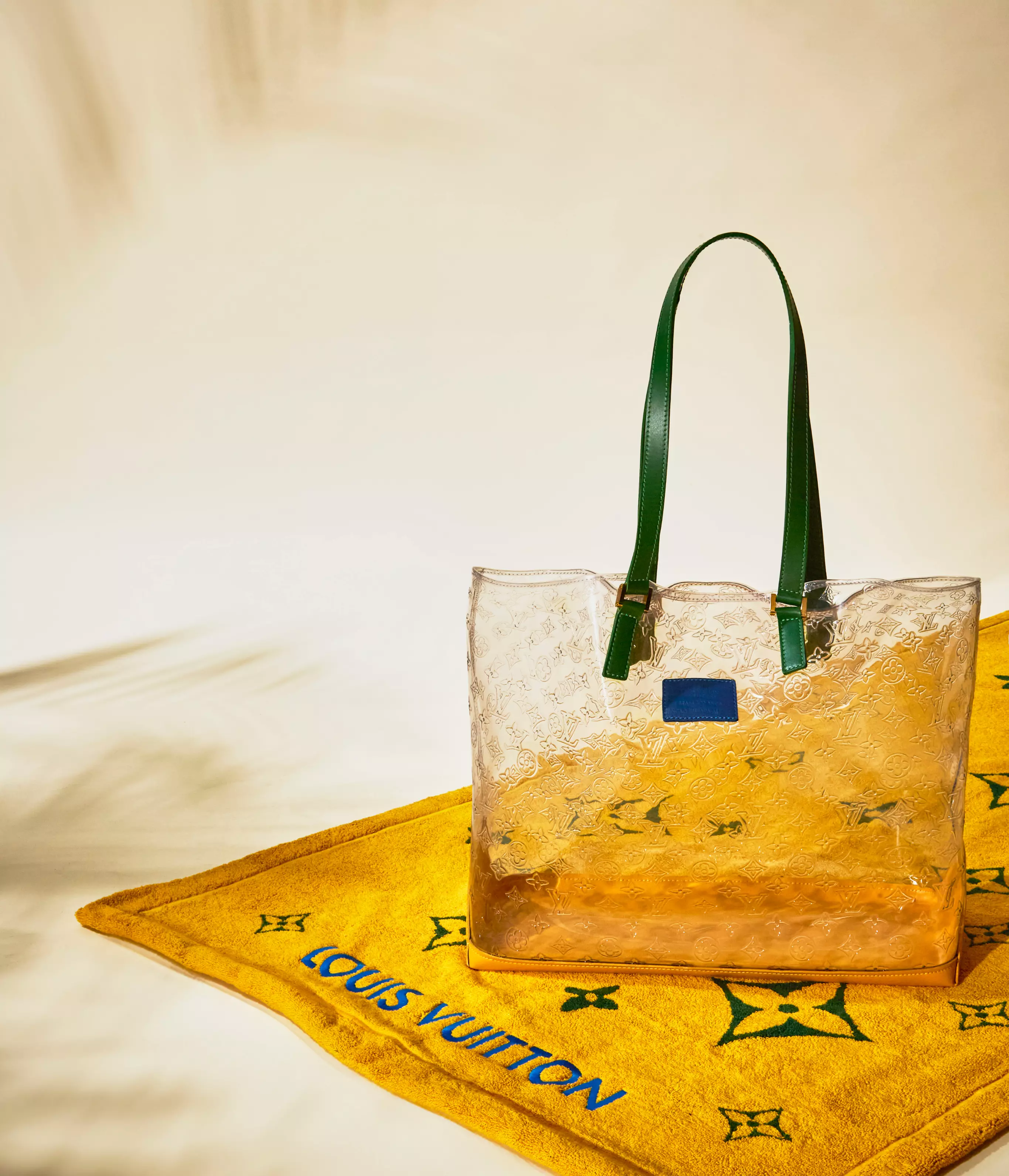 July Hero Banner: Louis Vuitton hero banner featuring a clear monogram tote with green handles displayed on a yellow Louis Vuitton towel against a cream backdrop.