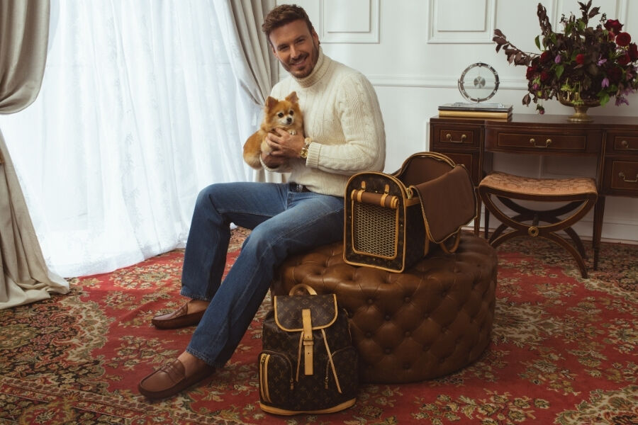 Shipping Policy: Male model in a white sweater and jeans holding a dog, sitting on a leather ottoman with a Louis Vuitton monogram canvas dog carrier and Louis Vuitton monogram Bosophere backpack.